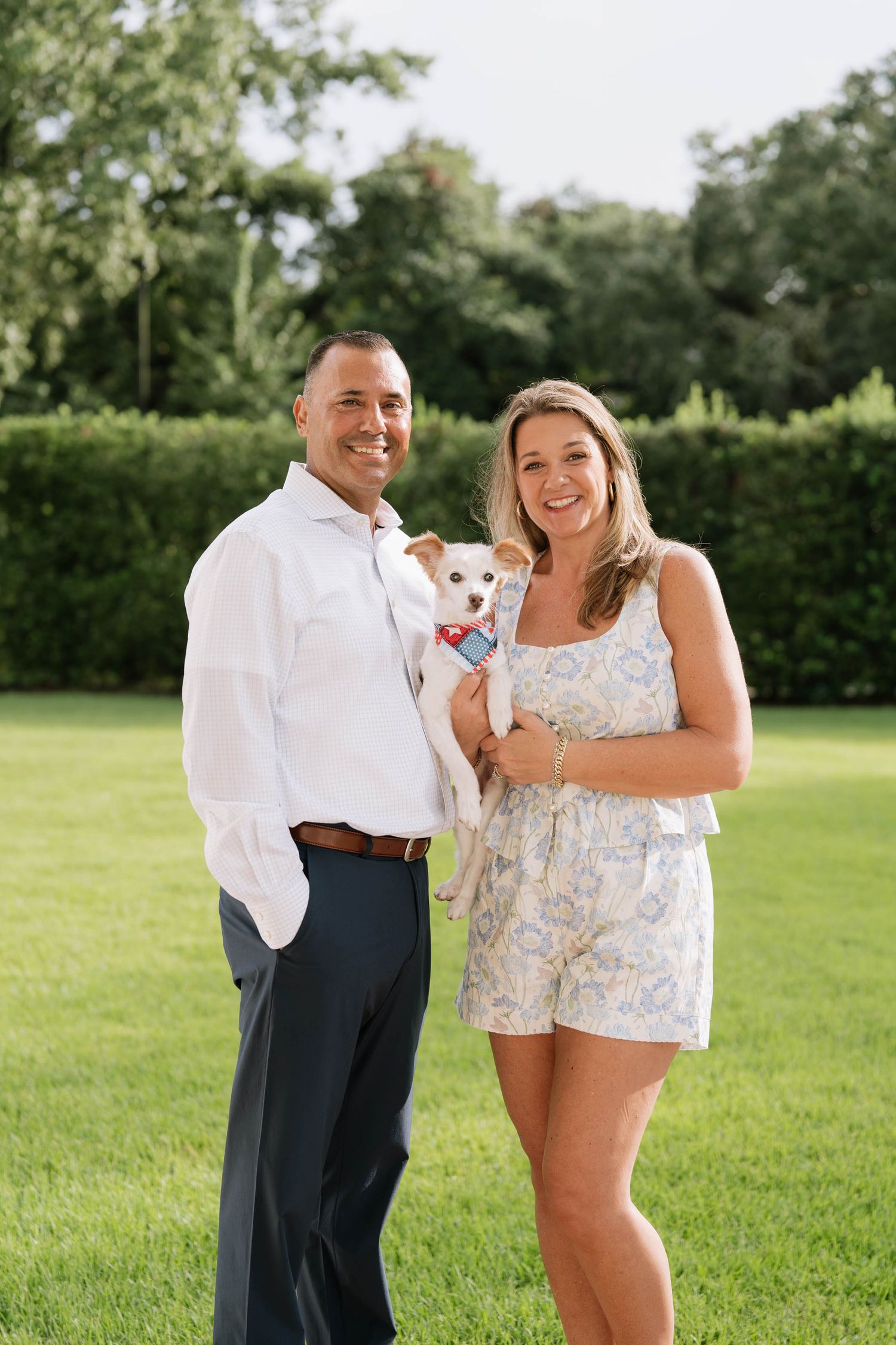 A couple poses with their small white dog on a sunny lawn with green trees in the background.