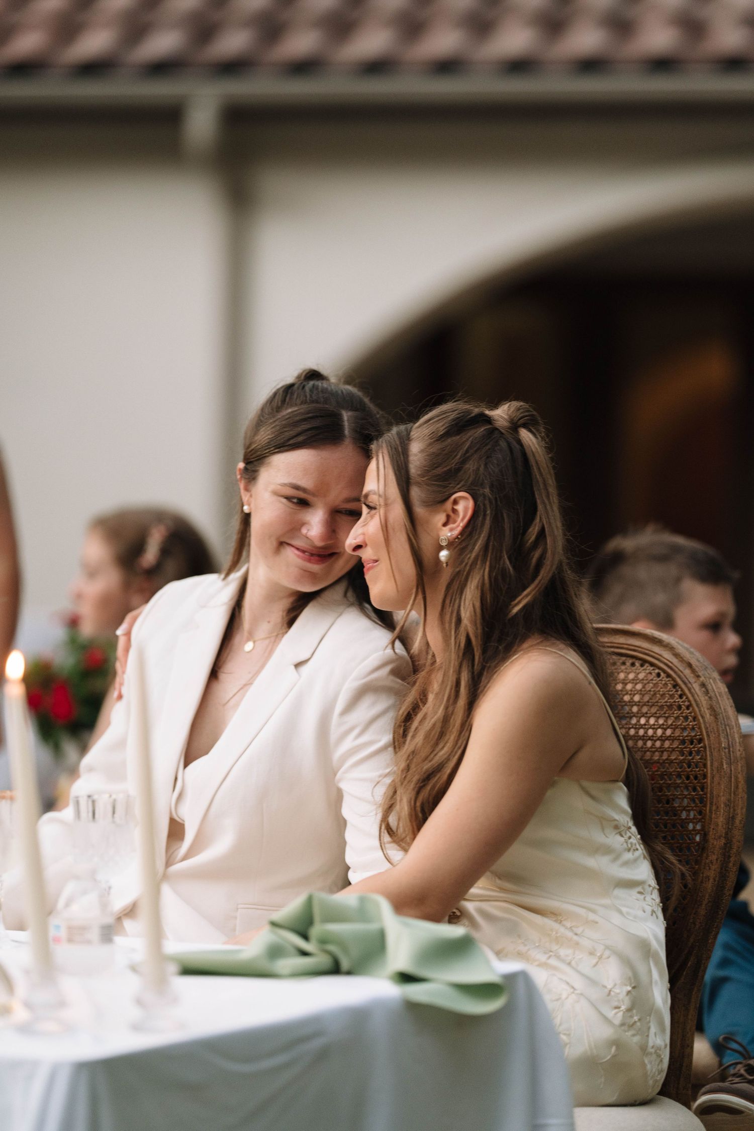 Two brides in white attire share an intimate moment at their wedding reception table outdoors.
