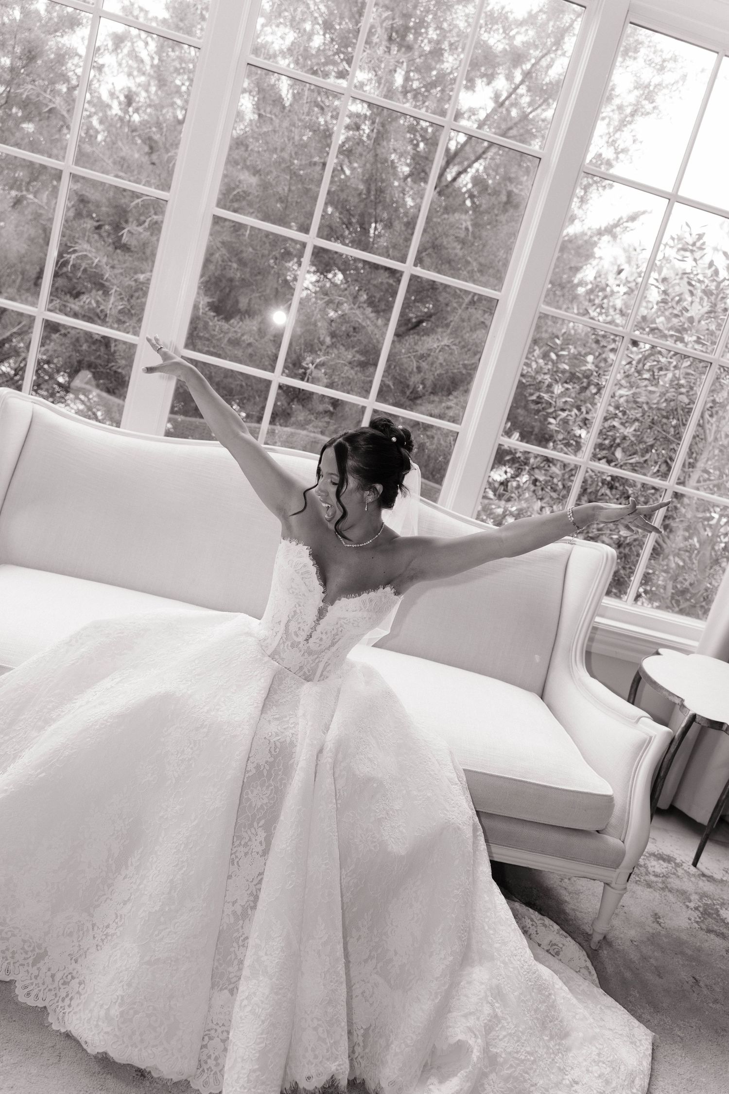 Black and white photo of a bride in a lace wedding gown sitting on a white sofa with arms outstretched.