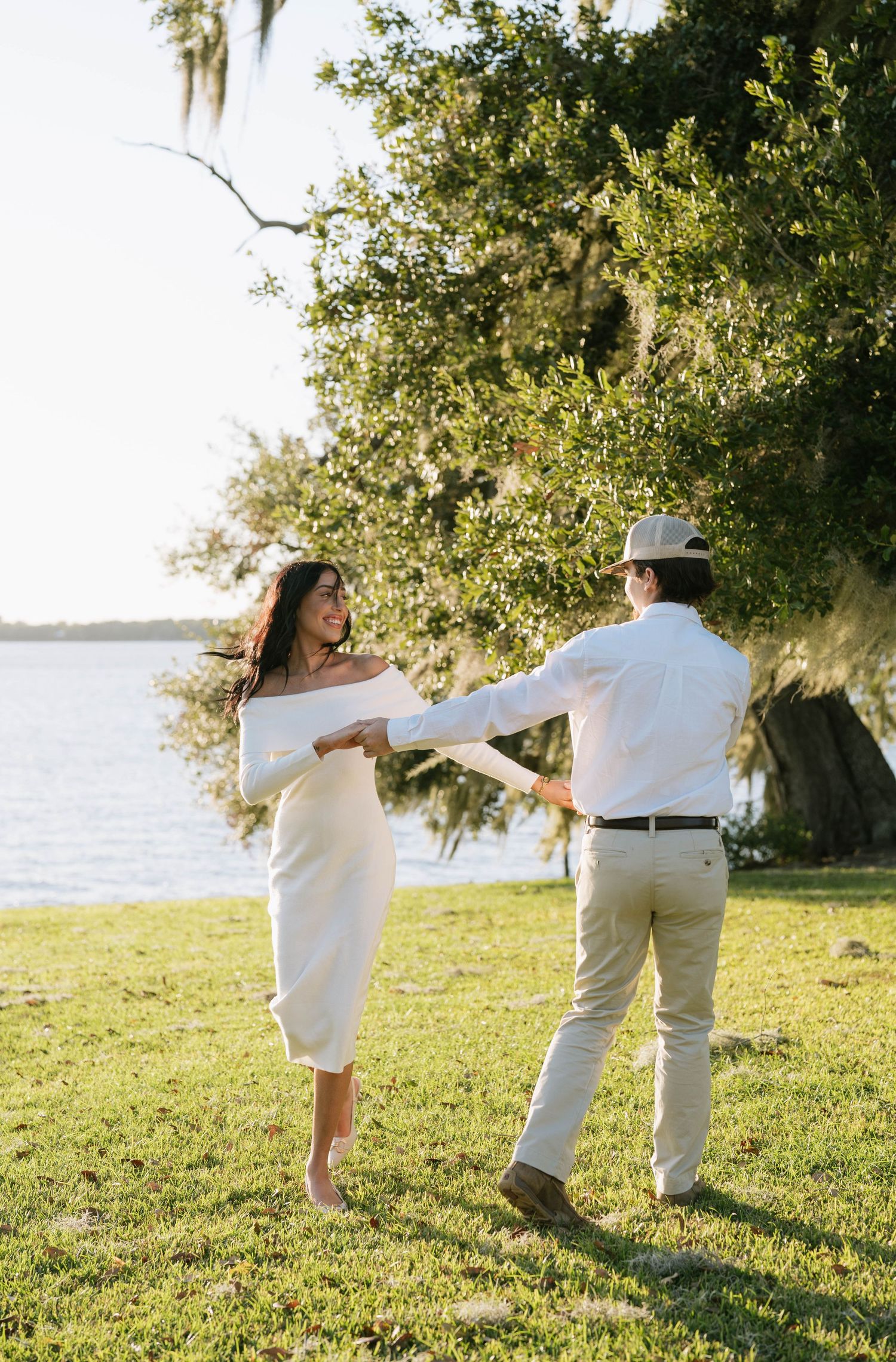 Couple in white outfits playfully walking on grass near waterfront.