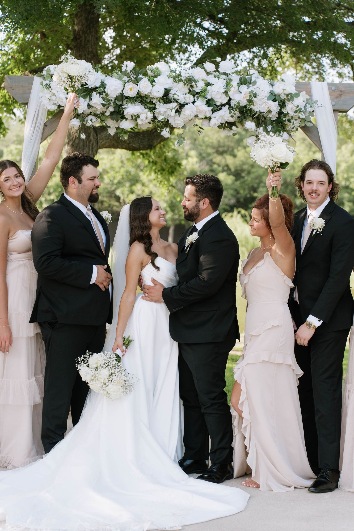 Wedding party celebrating outdoors under trees with joyful expressions.