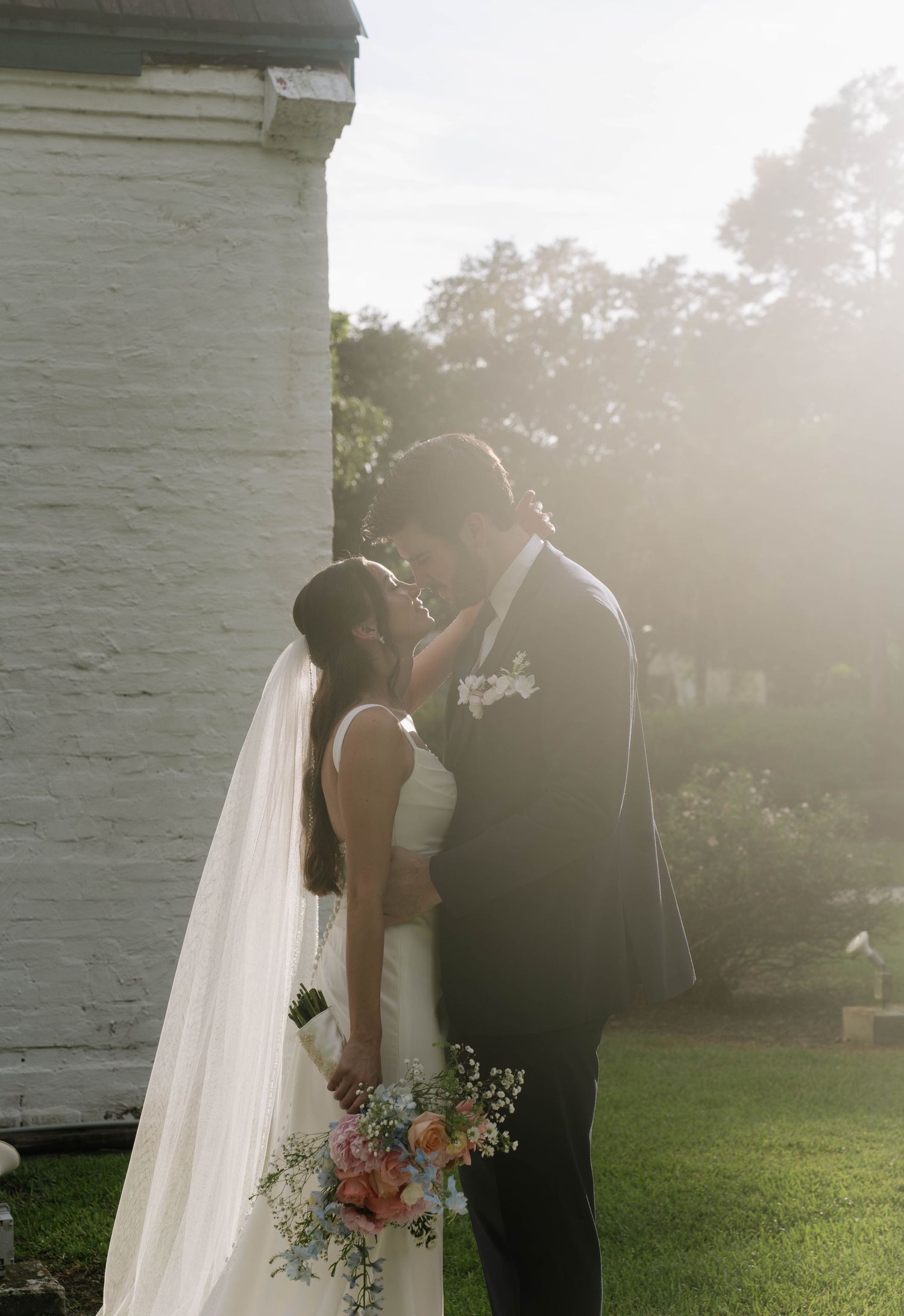 Wedding couple sharing an intimate moment in golden sunlight against a white brick wall.