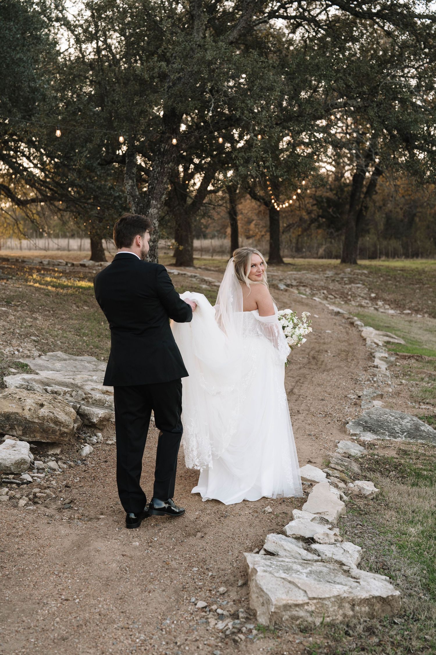 Groom in black tuxedo lifts bride's lace train as they walk a rustic stone path at golden hour.