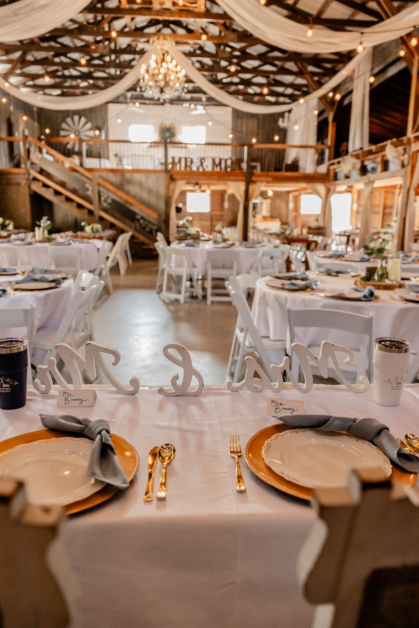 Elegant rustic barn wedding reception venue with white linens, gold chargers and romantic string light decor overhead.