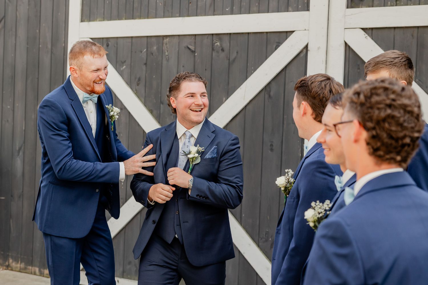Wedding party members in navy suits share candid laughs and conversations in front of a dark barn wall.