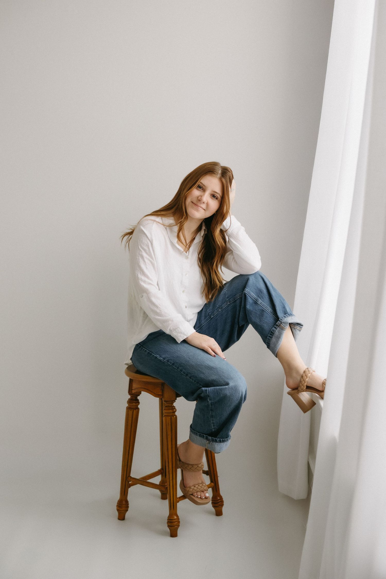 A person in casual attire with a white shirt and jeans sits relaxed on a wooden stool near a window.
