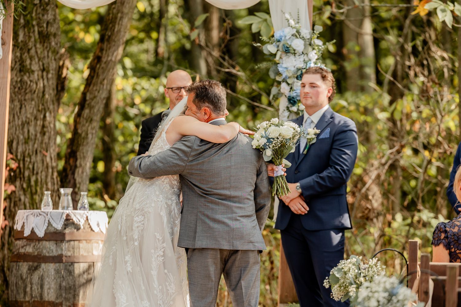 Wedding ceremony taking place outdoors under a white arch decorated with flowers in a wooded setting.