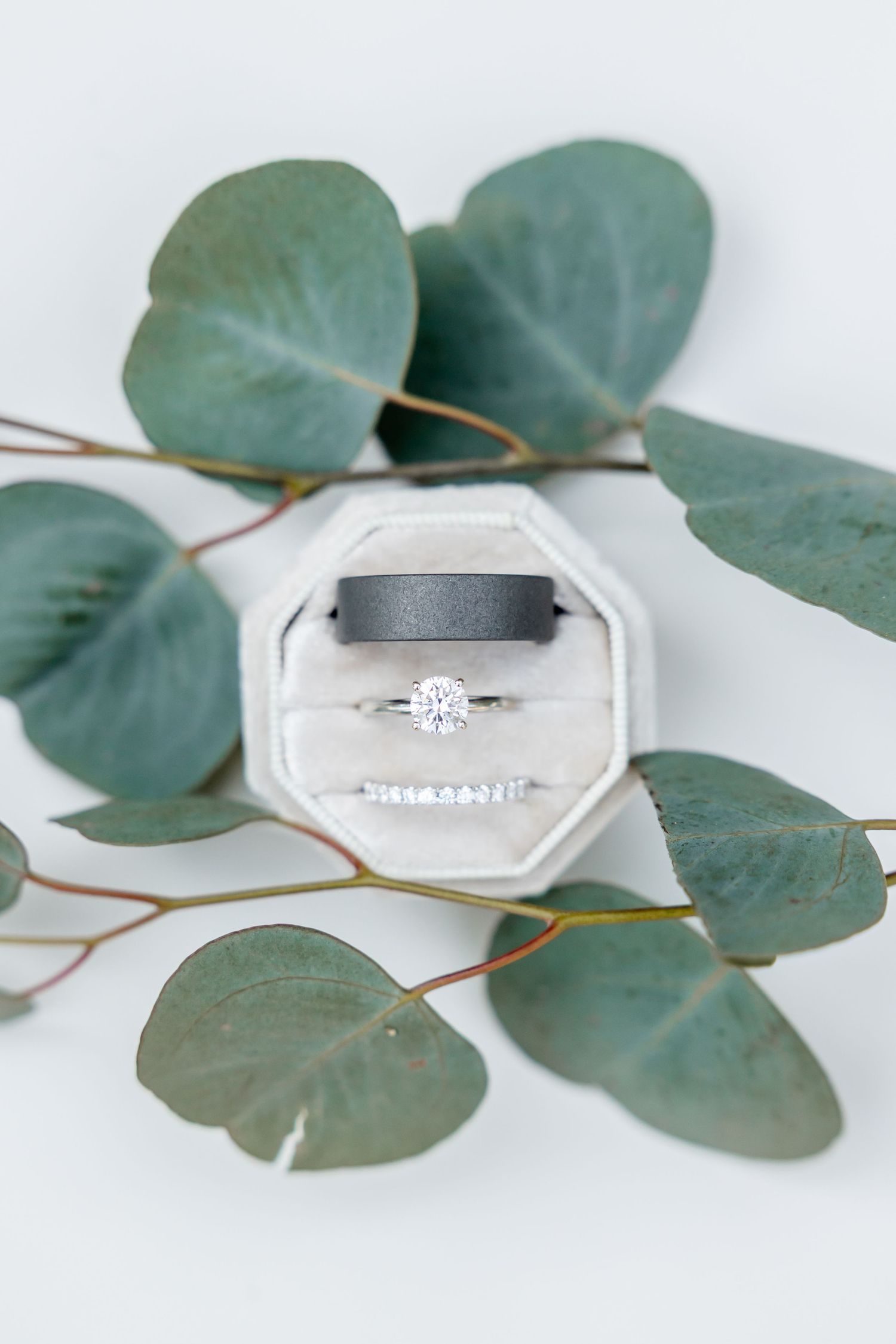Elegant engagement rings and wedding bands displayed with soft eucalyptus leaves on white marble surface.