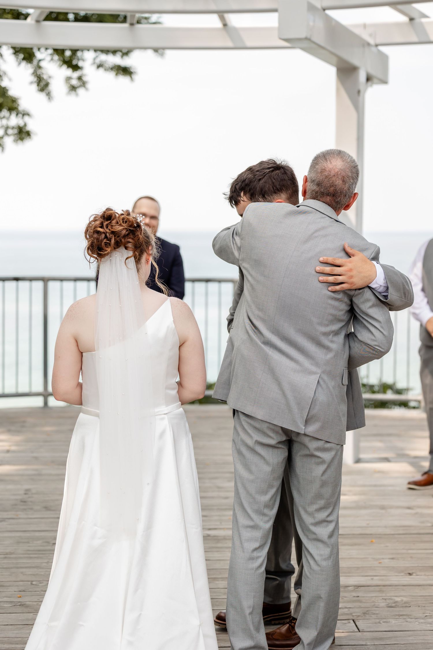 Wedding ceremony participants share emotional moments on a waterfront deck under white pergola.