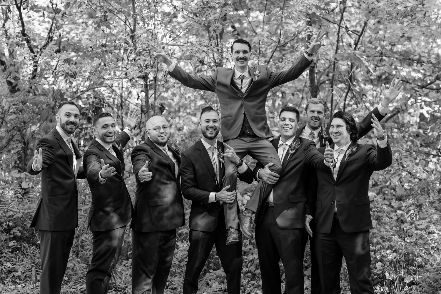 Group of groomsmen in dark suits striking fun poses against autumn trees.