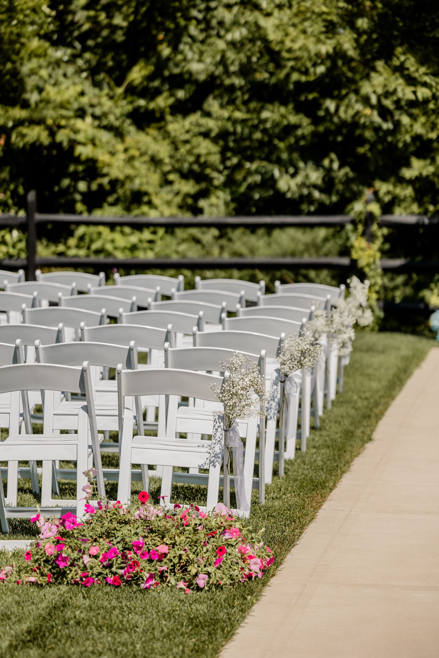 White wedding chairs arranged in rows with bright pink flowers lining the outdoor ceremony aisle.