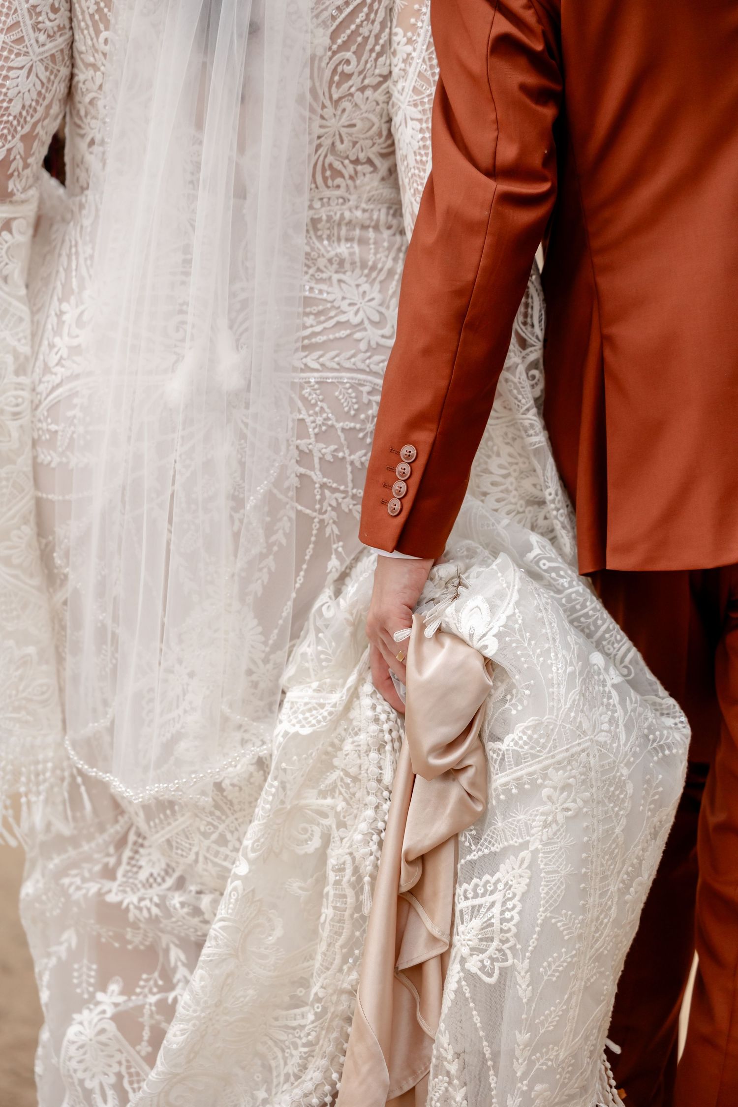 Close-up detail of rust-colored suit jacket against white lace wedding dress.