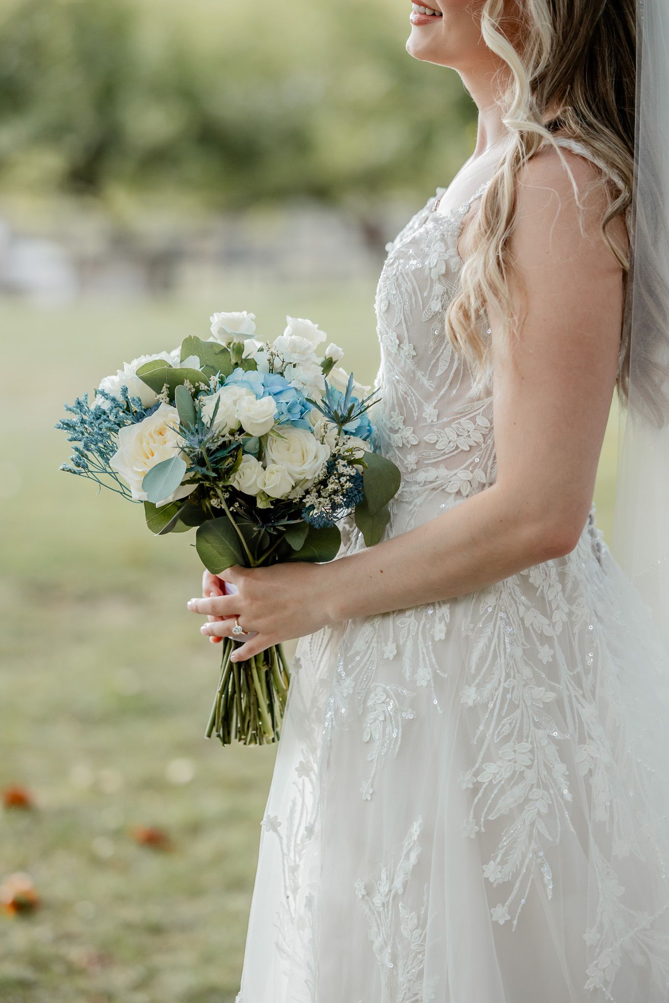 A bride in a delicate lace wedding dress holds a beautiful bouquet of white and blue flowers against a soft outdoor backdrop.