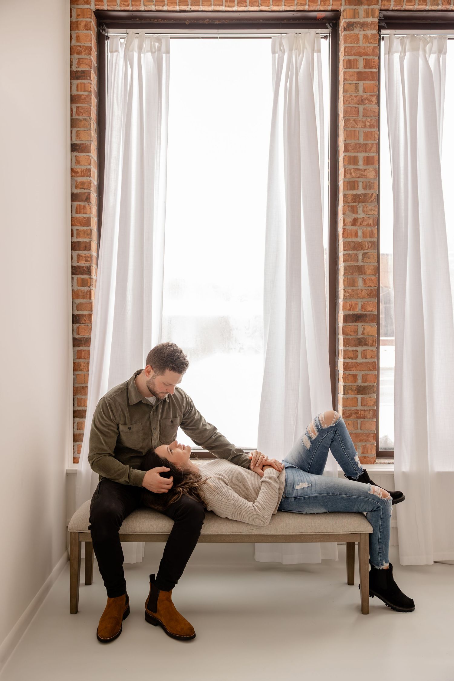 A person in casual attire sits on a windowsill in a bright room with exposed brick walls and white curtains.
