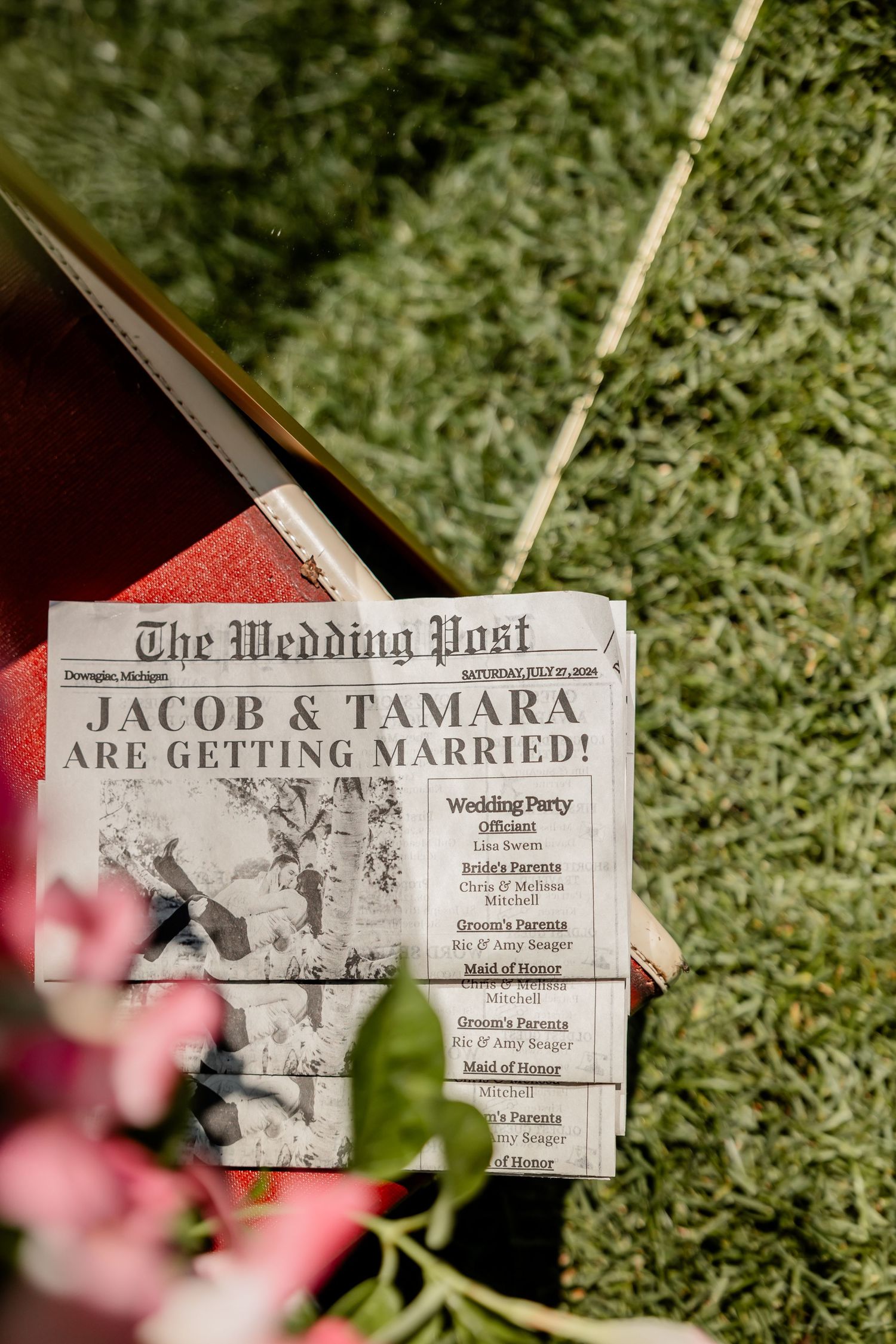 Vintage newspaper style wedding announcements displayed on grass with pink flowers.
