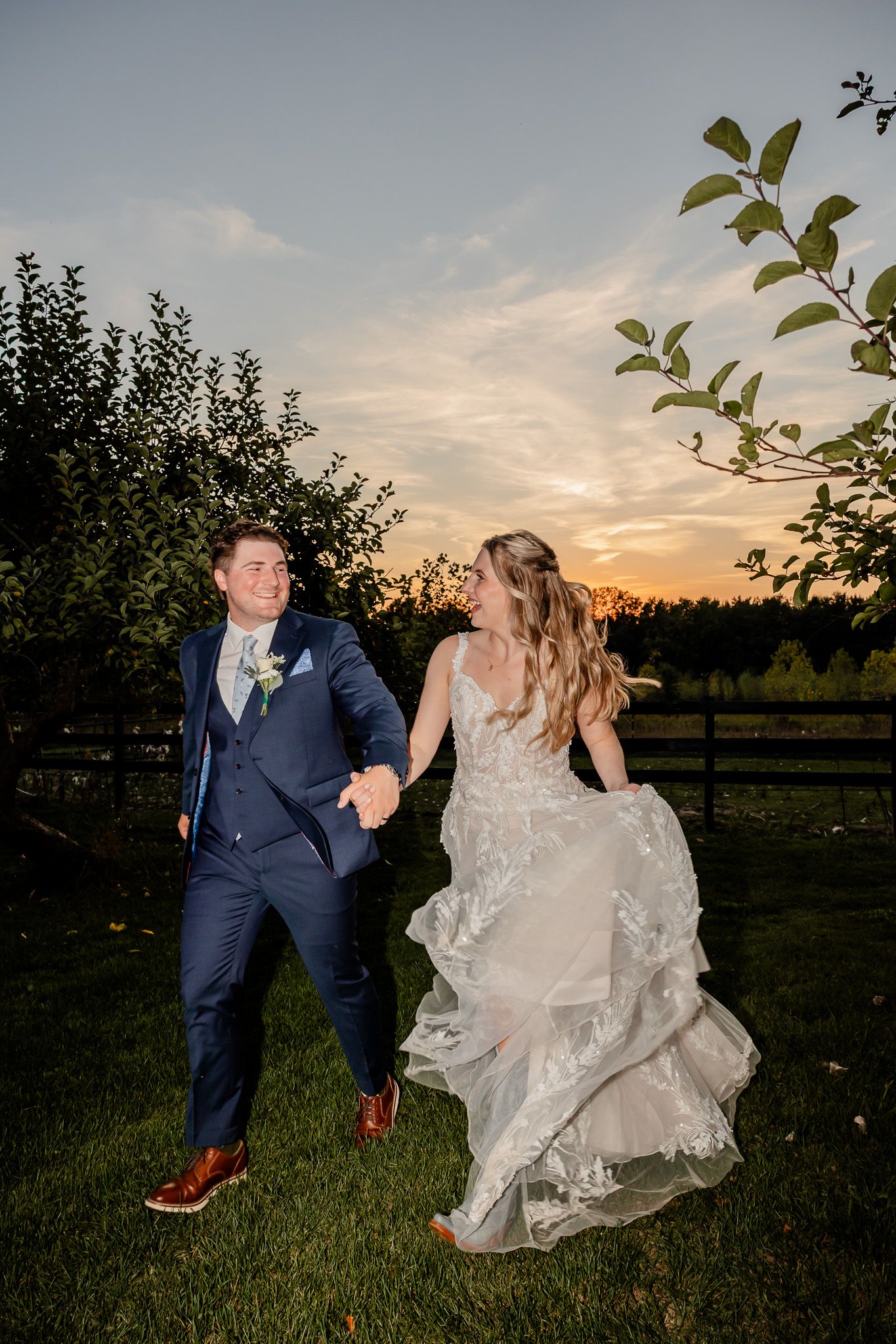 A couple in wedding attire dances playfully in a grassy field at sunset.