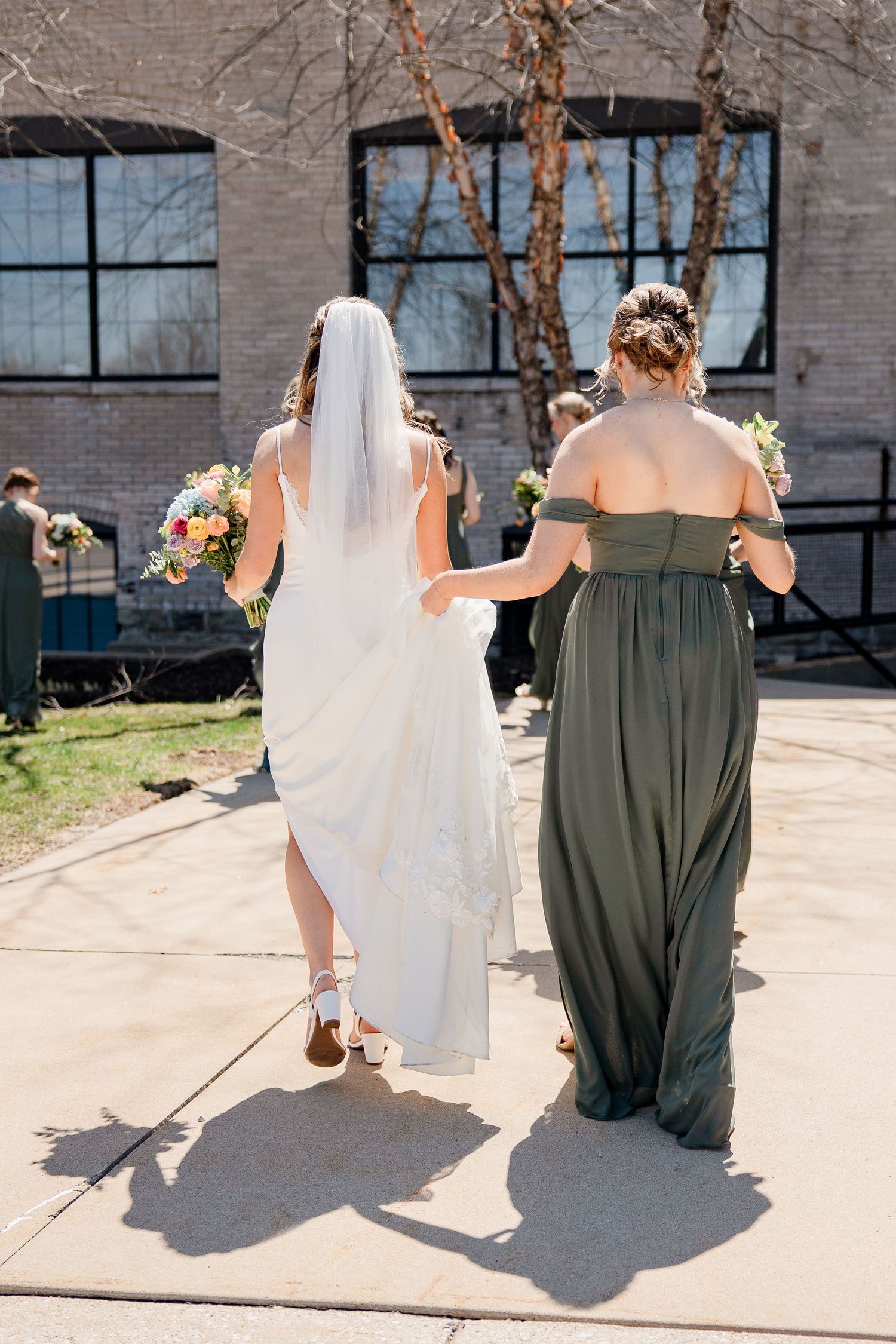 Bride and bridesmaids in olive green dresses walking outside a brick building on a sunny day.