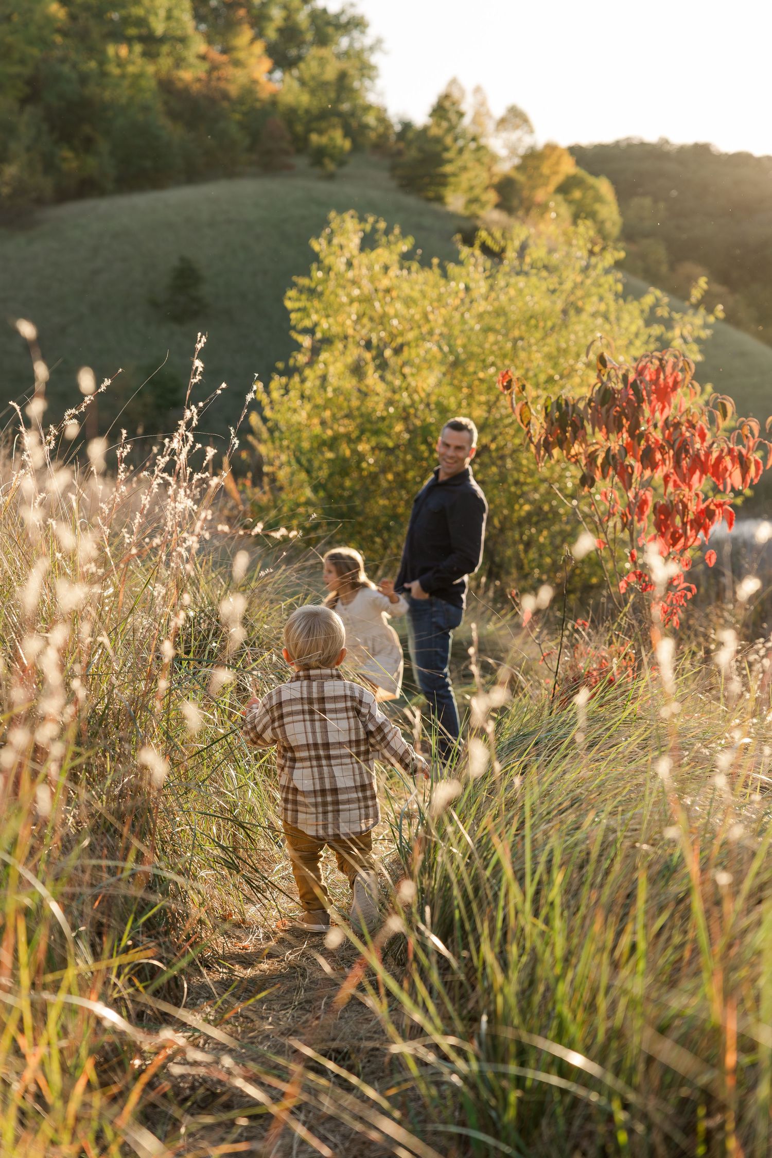 Family walking through golden autumn meadow with lush green hills and red foliage in warm sunset light.