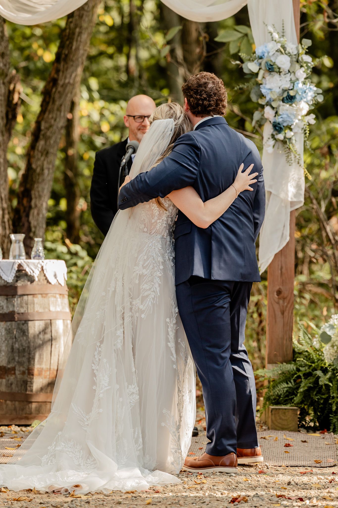 A bride and groom share their first kiss at a rustic outdoor wedding ceremony decorated with white draping and flowers.