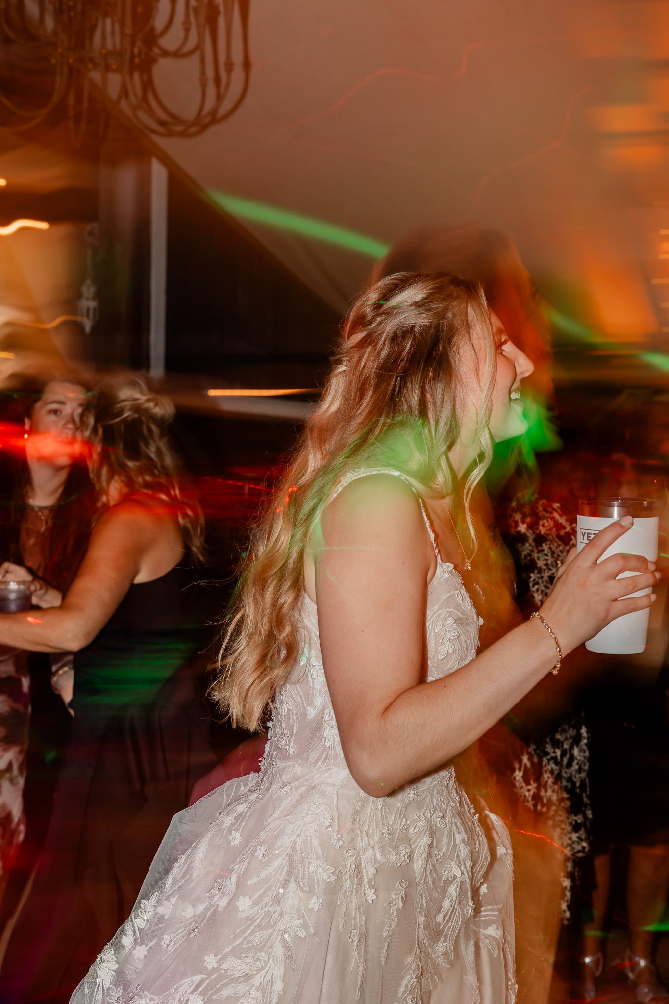 Motion-blurred dancers in white dresses celebrate at a wedding reception with colorful lights creating a dynamic atmosphere.