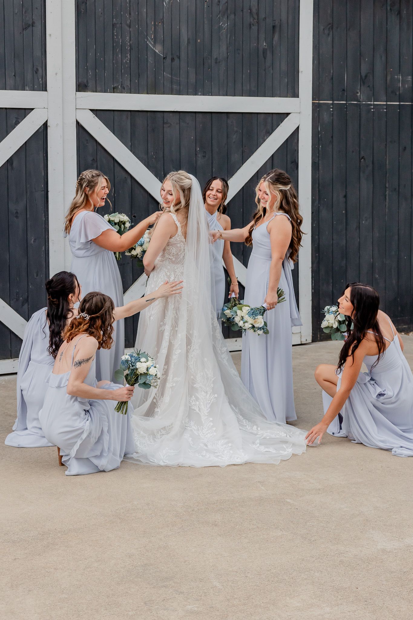 Bridesmaids in light blue dresses arrange a bride's long train against a dark barn wall backdrop.