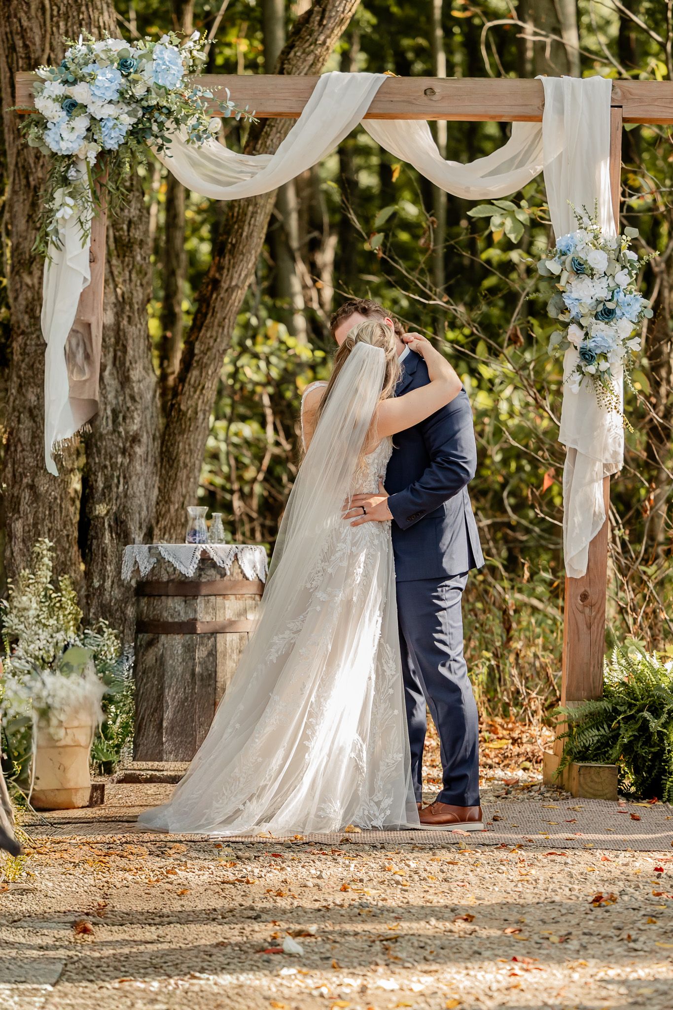 A couple shares a romantic embrace under white fabric drapes and trees at an outdoor wedding venue.