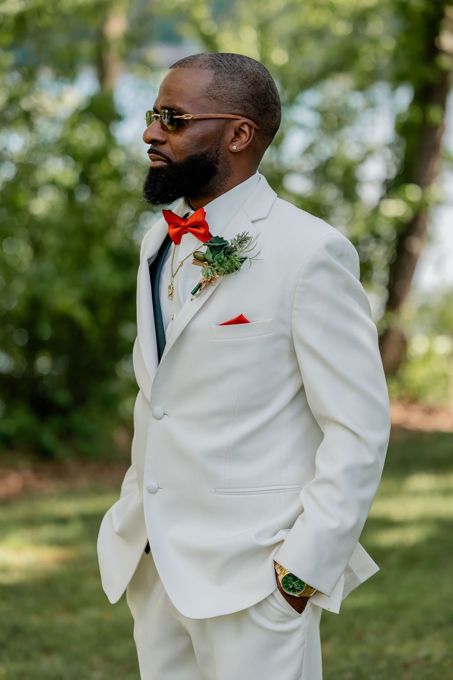 Stylish groom in white suit with red bowtie and pocket square posing outdoors.