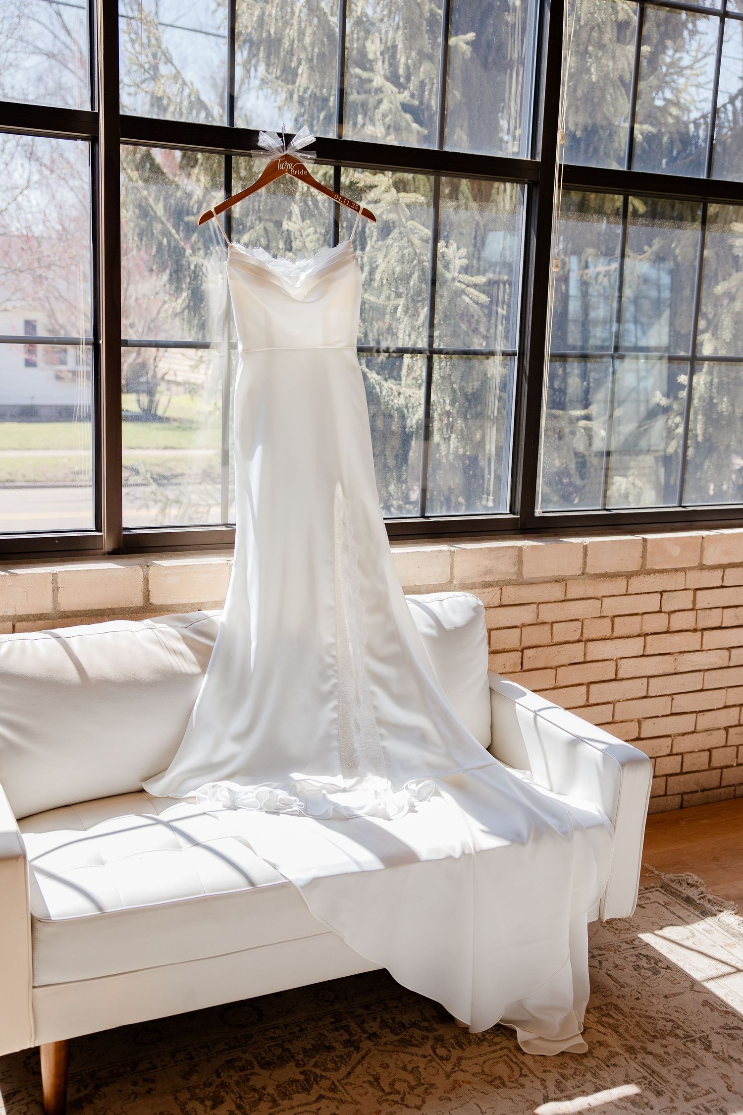 White wedding dresses hang elegantly by windows in a sunlit brick room with cascading natural light.