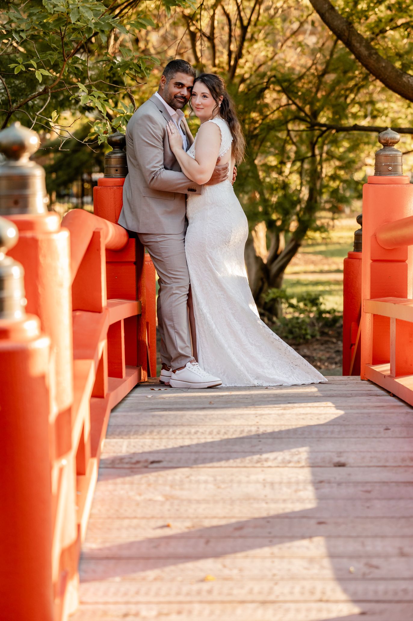 A couple in wedding attire embraces on a rustic bridge with orange railings during a sunset photoshoot.