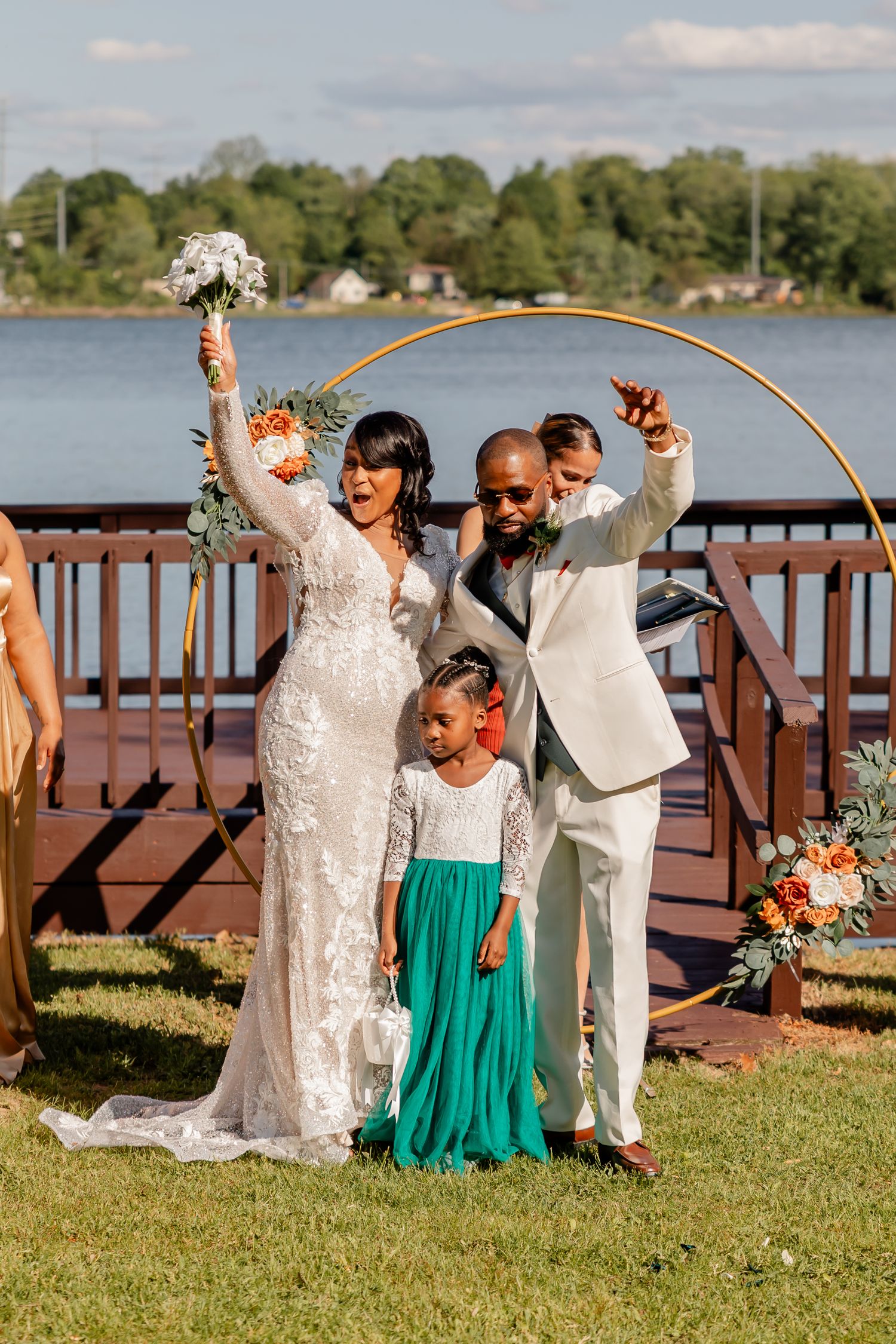 Wedding celebration under golden arch by lakeside with couple and child in teal dress.