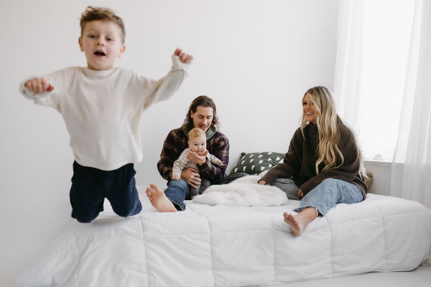 A family plays together on a white bed in a bright sunlit room, creating joyful moments at home.