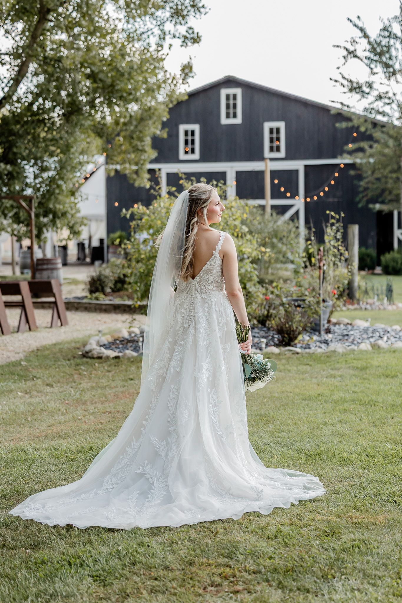 A flowing white wedding dress with a long train and veil photographed in a garden setting with dark barns in the background.