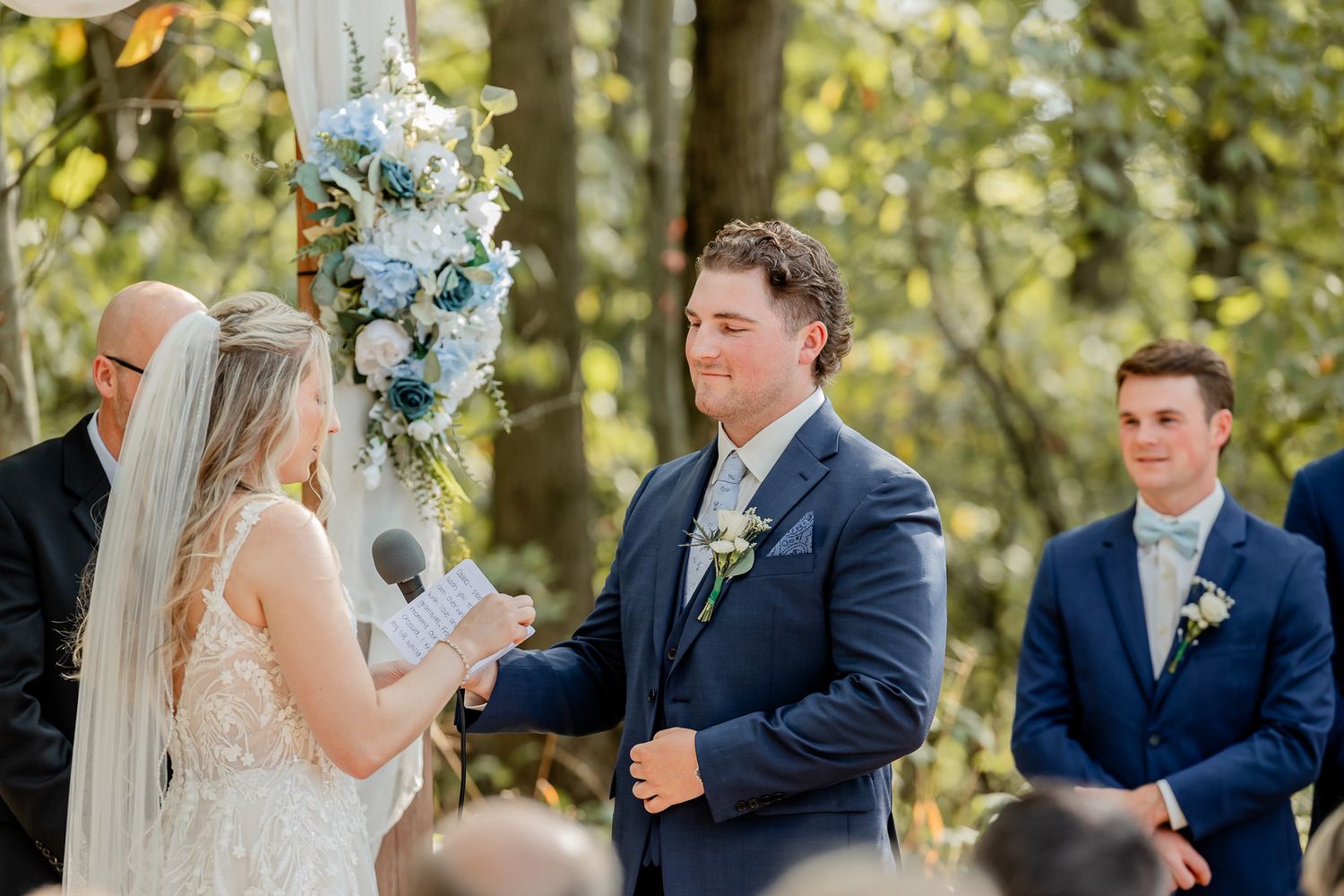 A wedding ceremony takes place outdoors with blue floral decorations and navy attire against a natural wooded backdrop.