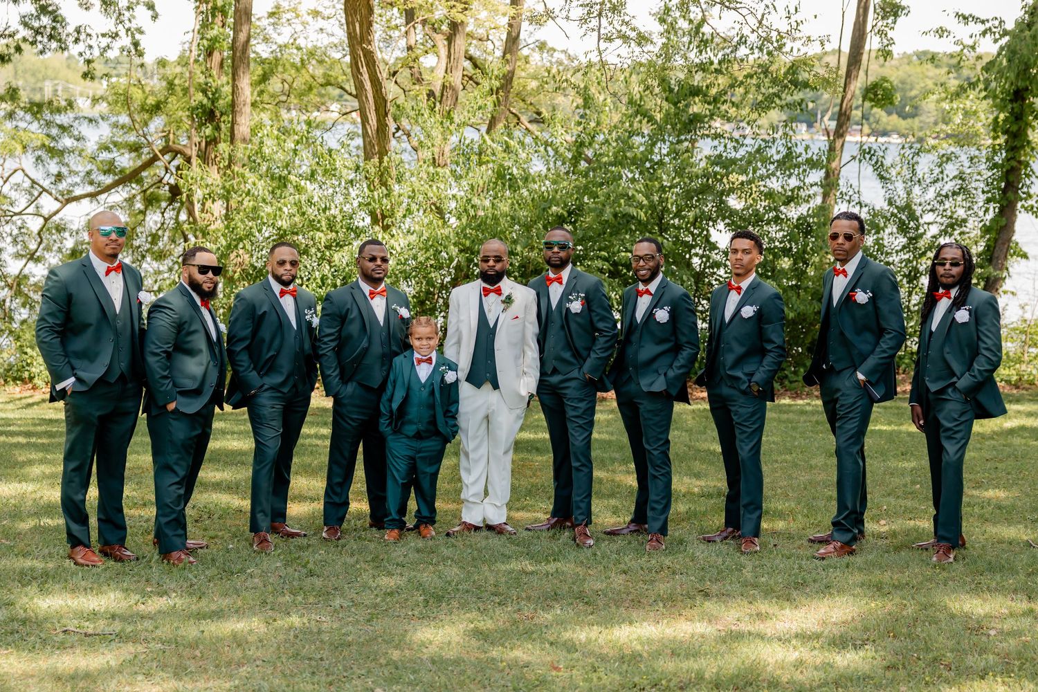Wedding party in coordinated navy suits and one white suit pose together on grassy lawn.
