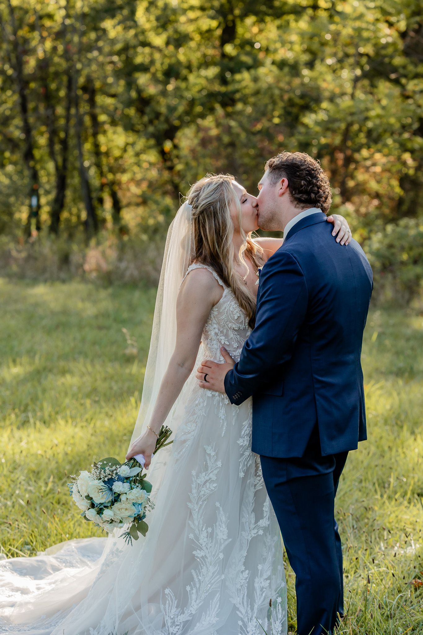 A couple shares a romantic kiss in a sunlit meadow surrounded by trees while wearing wedding attire.