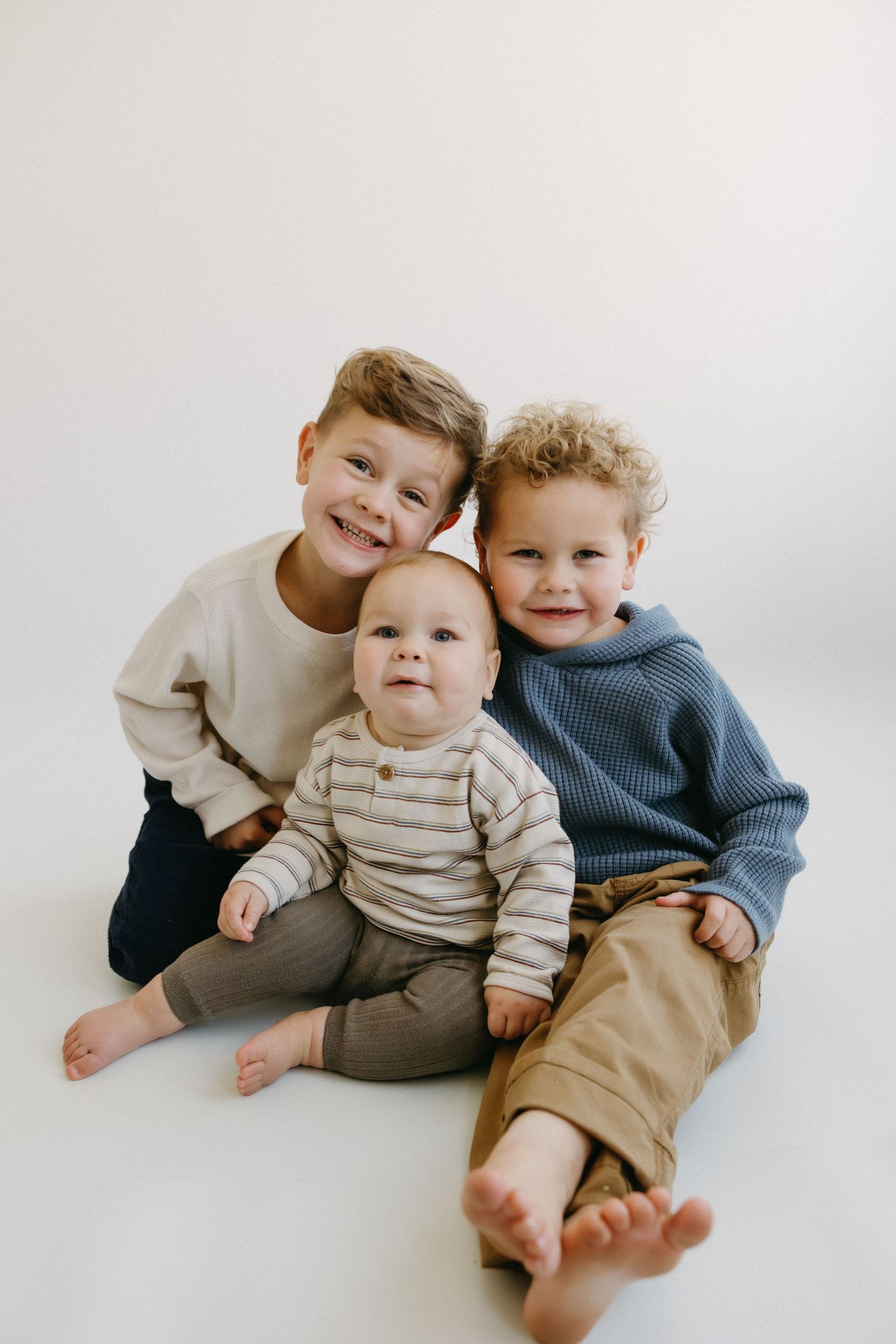 Three young children in cozy sweaters sit together on a white background, smiling and posing casually.