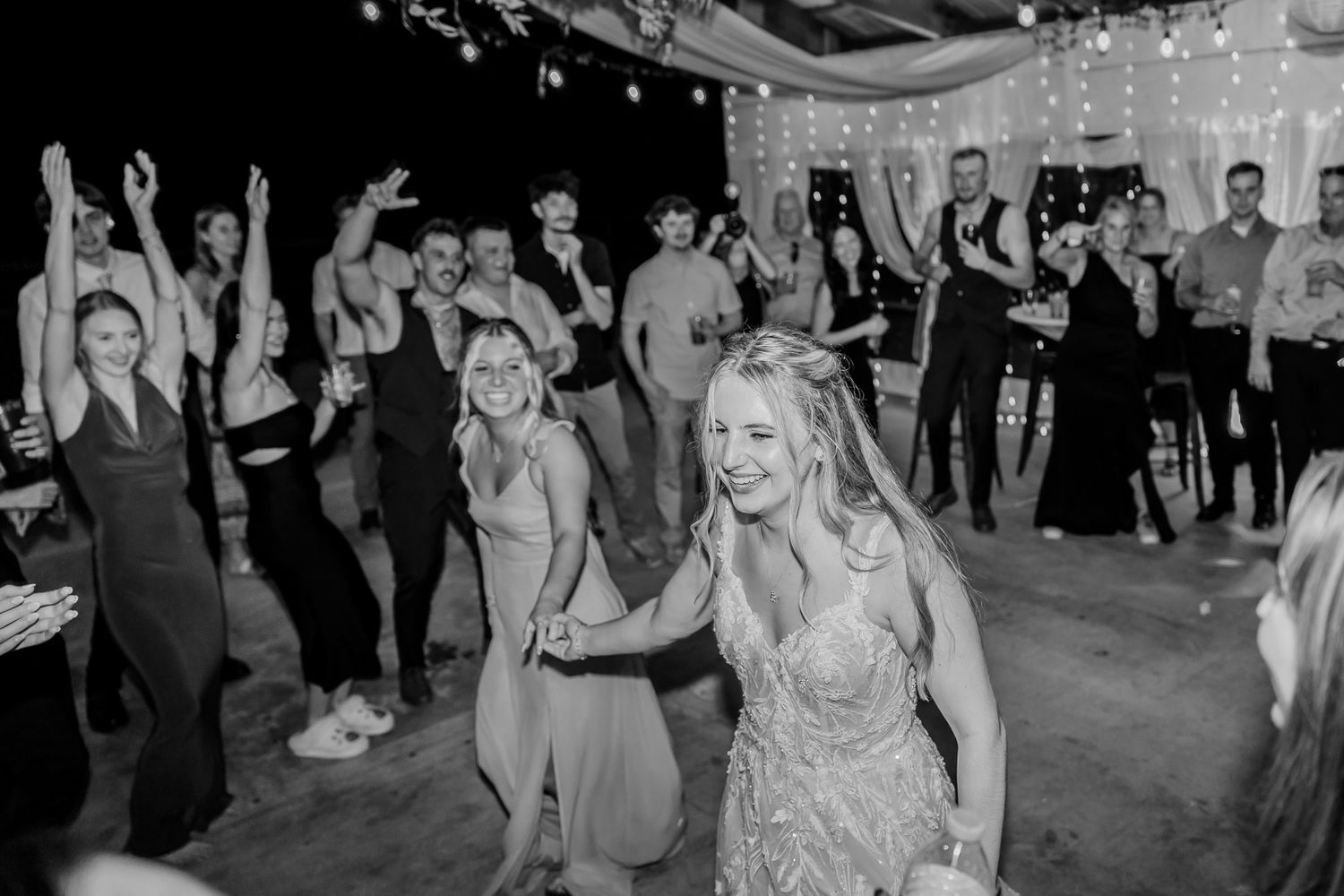 Black and white photo of people dancing and celebrating at a party with string lights overhead.