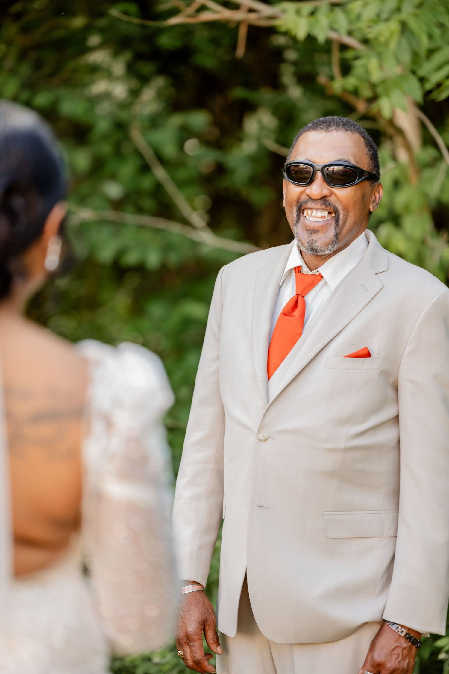 Wedding guest wearing a light beige suit with orange tie and sunglasses smiles joyfully outdoors.