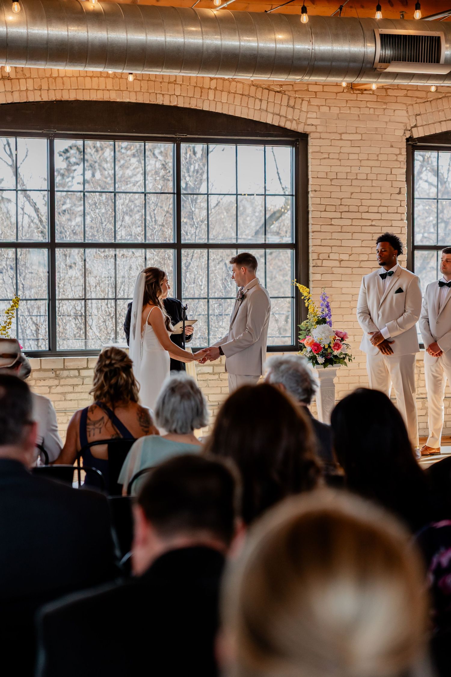 Indoor wedding ceremony in industrial venue with exposed brick walls and large windows.
