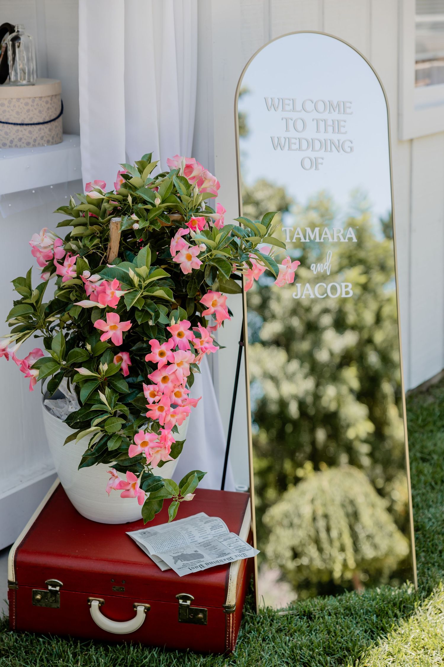 Wedding welcome sign with pink flowers in red vintage suitcase on grass.