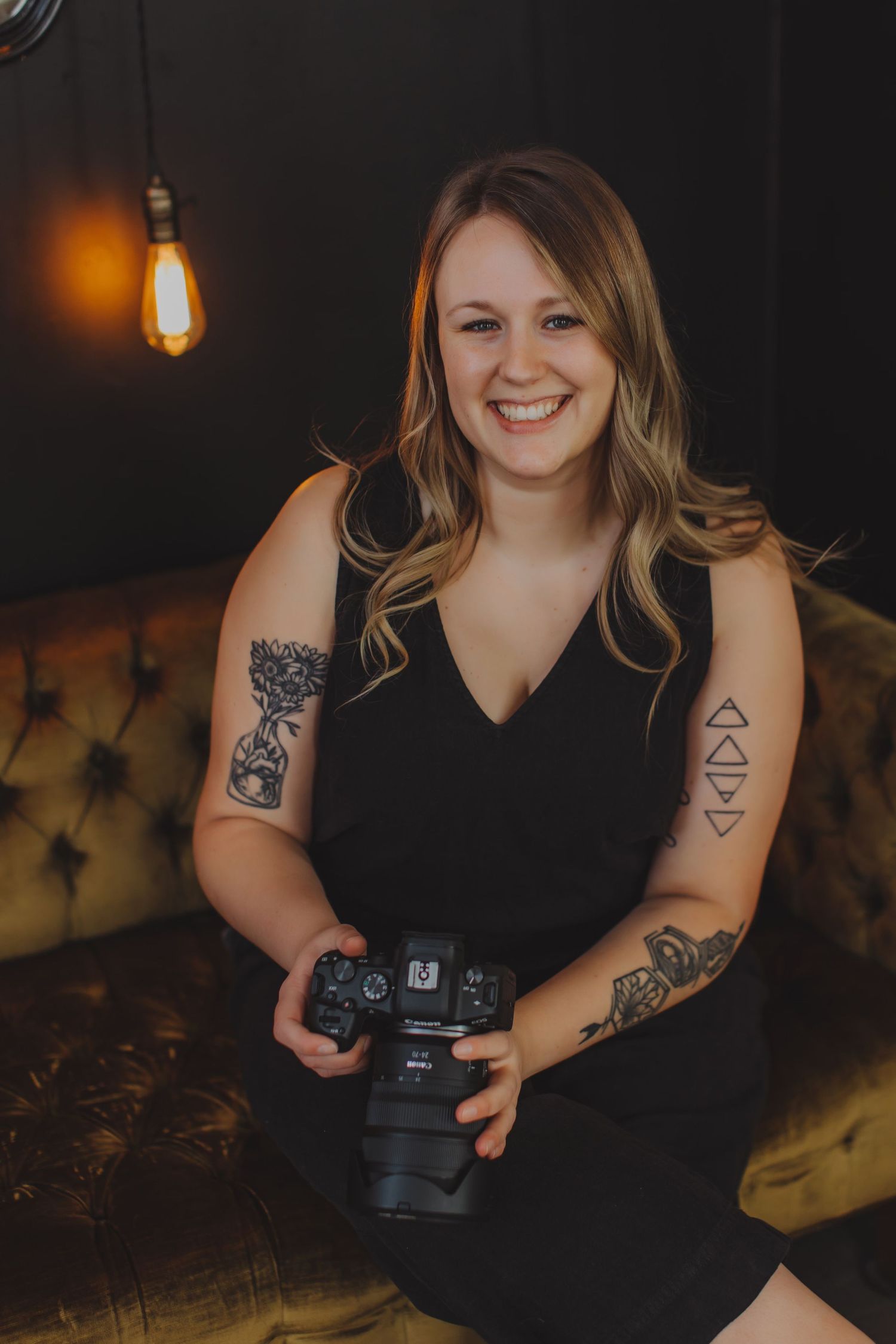 Photographer in black dress with tattoos holds camera in dimly lit setting.