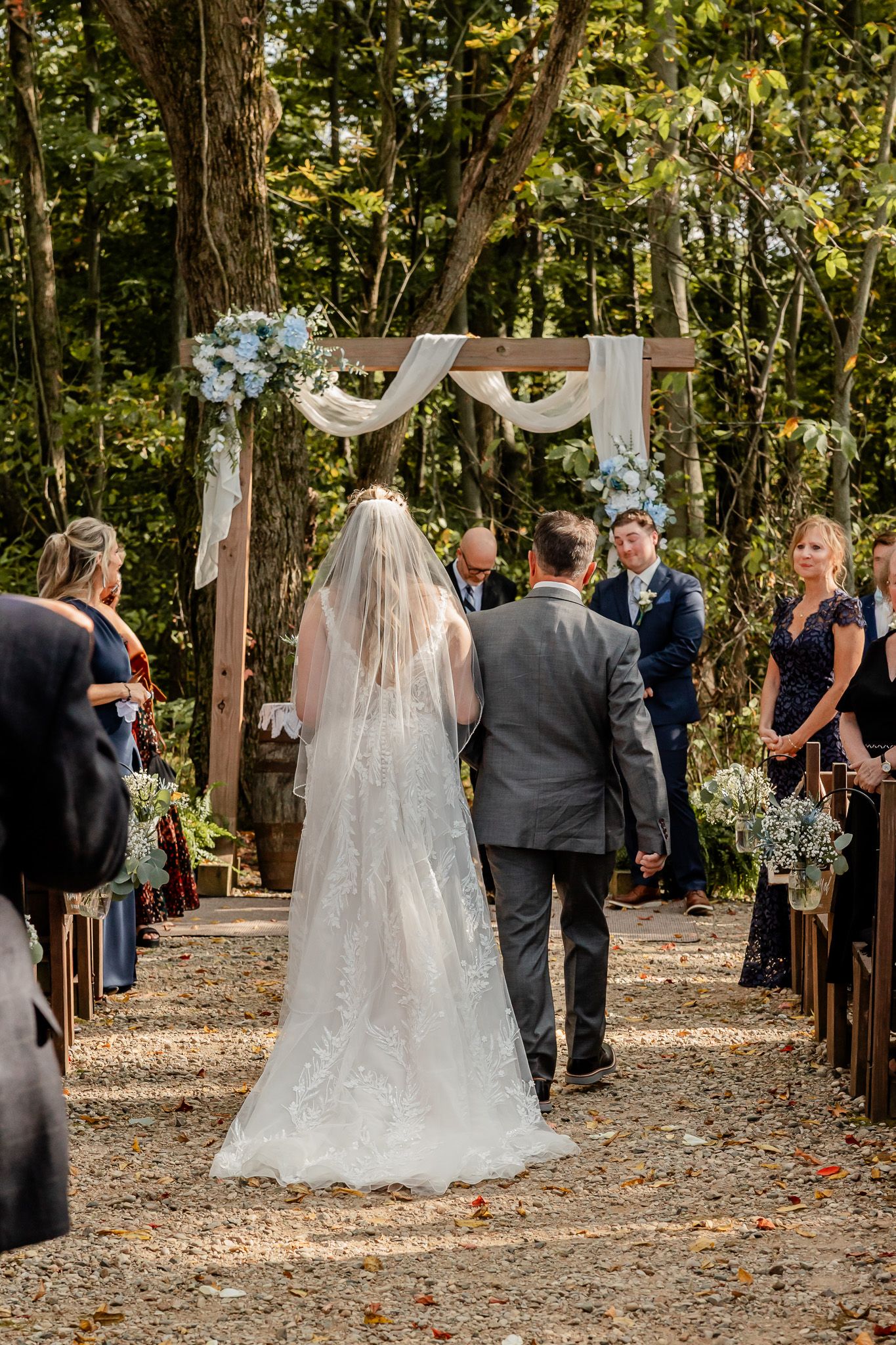 Outdoor woodland wedding ceremony with rustic wooden arch decorated with white fabric and flowers on a natural forest path.