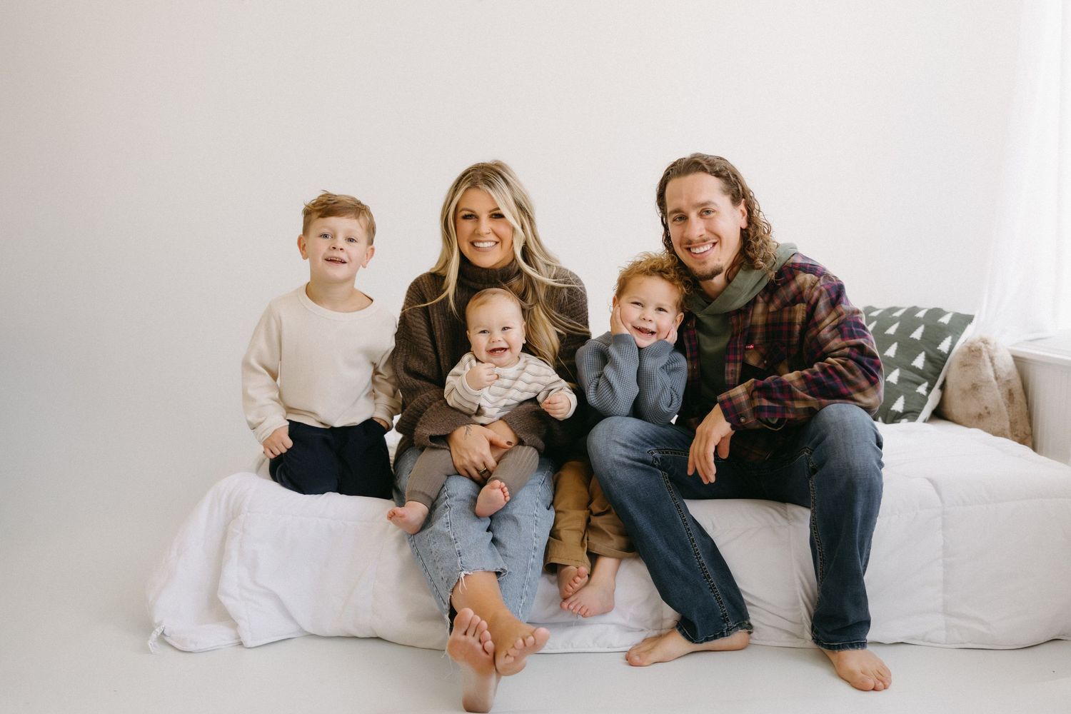 Family sits together on white couch wearing coordinating neutral colored outfits for casual portrait session.