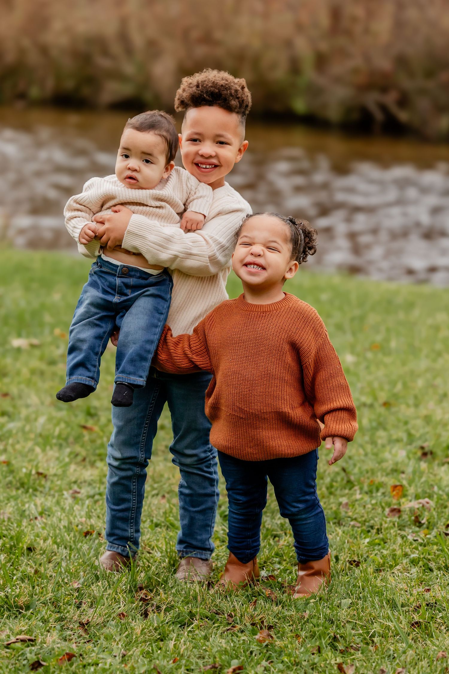 Three young children pose outdoors near a stream in fall, wearing cozy sweaters and jeans, smiling warmly.
