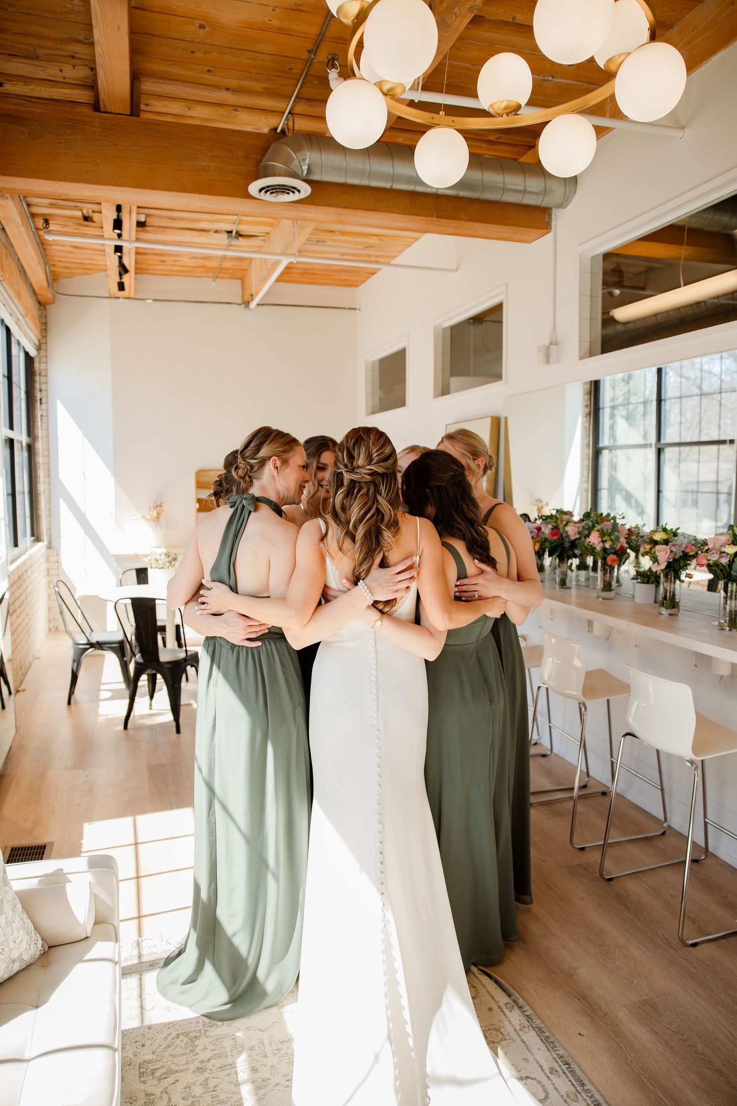 Wedding party in sage green and white dresses huddles together in venue with wooden ceiling and paper lanterns.