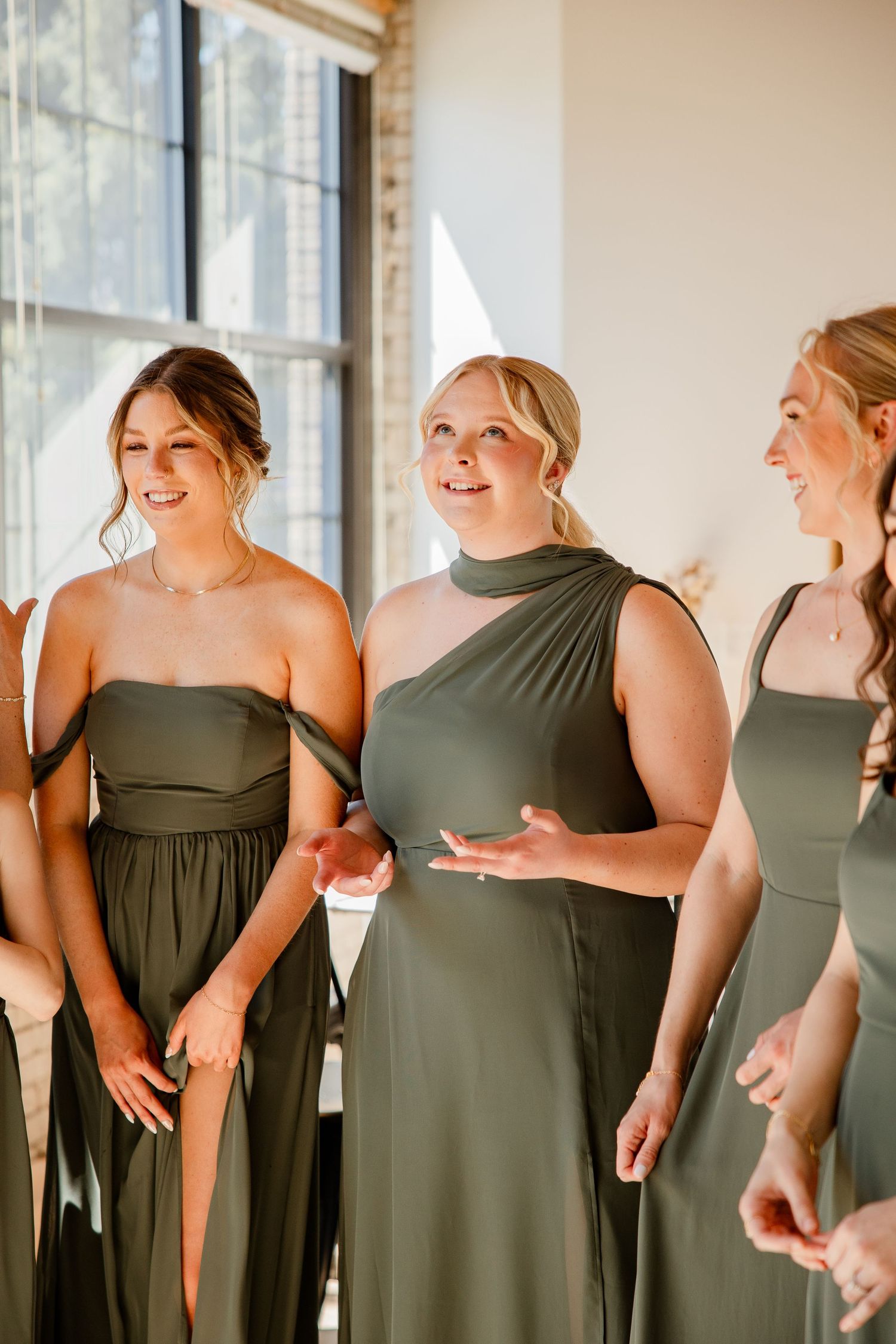Bridesmaids in olive green dresses share candid moments in bright sunlit room.