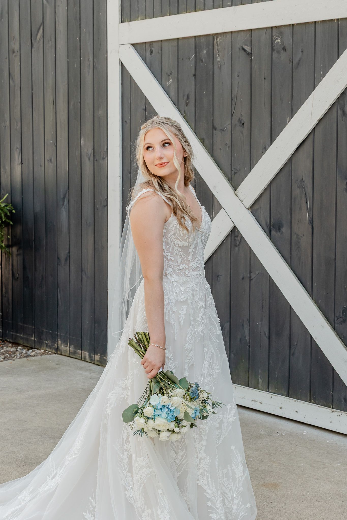 A white lace wedding dress with a flowing veil is showcased against a dark barn wall with white trim details.