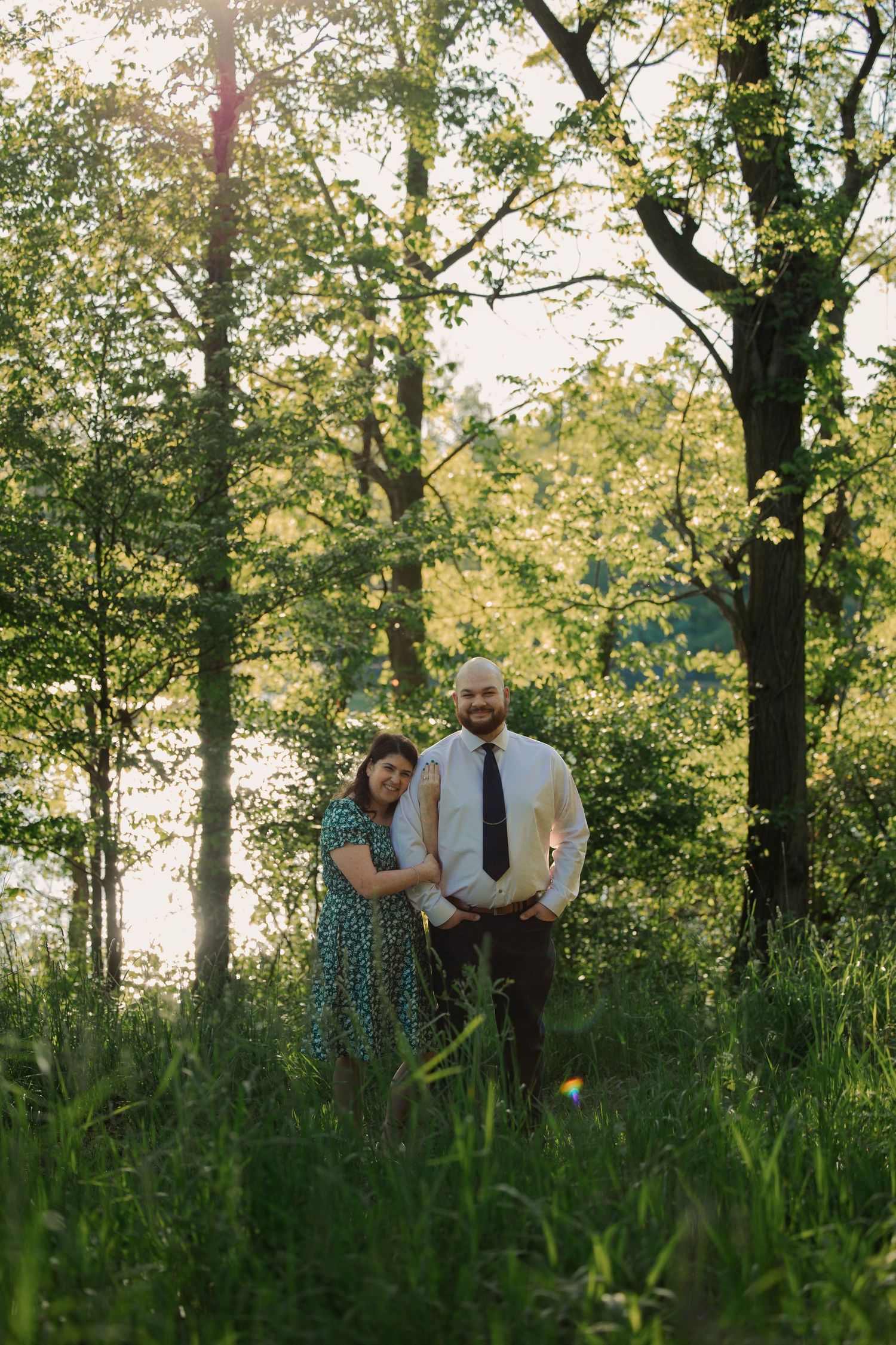 Couple poses together in tall grass with sunlight filtering through trees behind them. Dowagiac