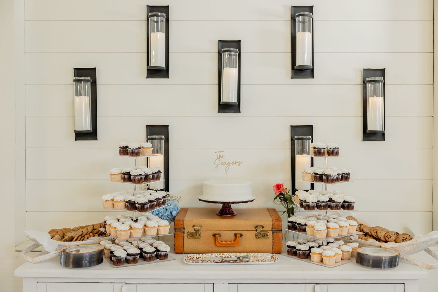 Wedding dessert display with wall sconces and decorative treats arranged on a white backdrop.