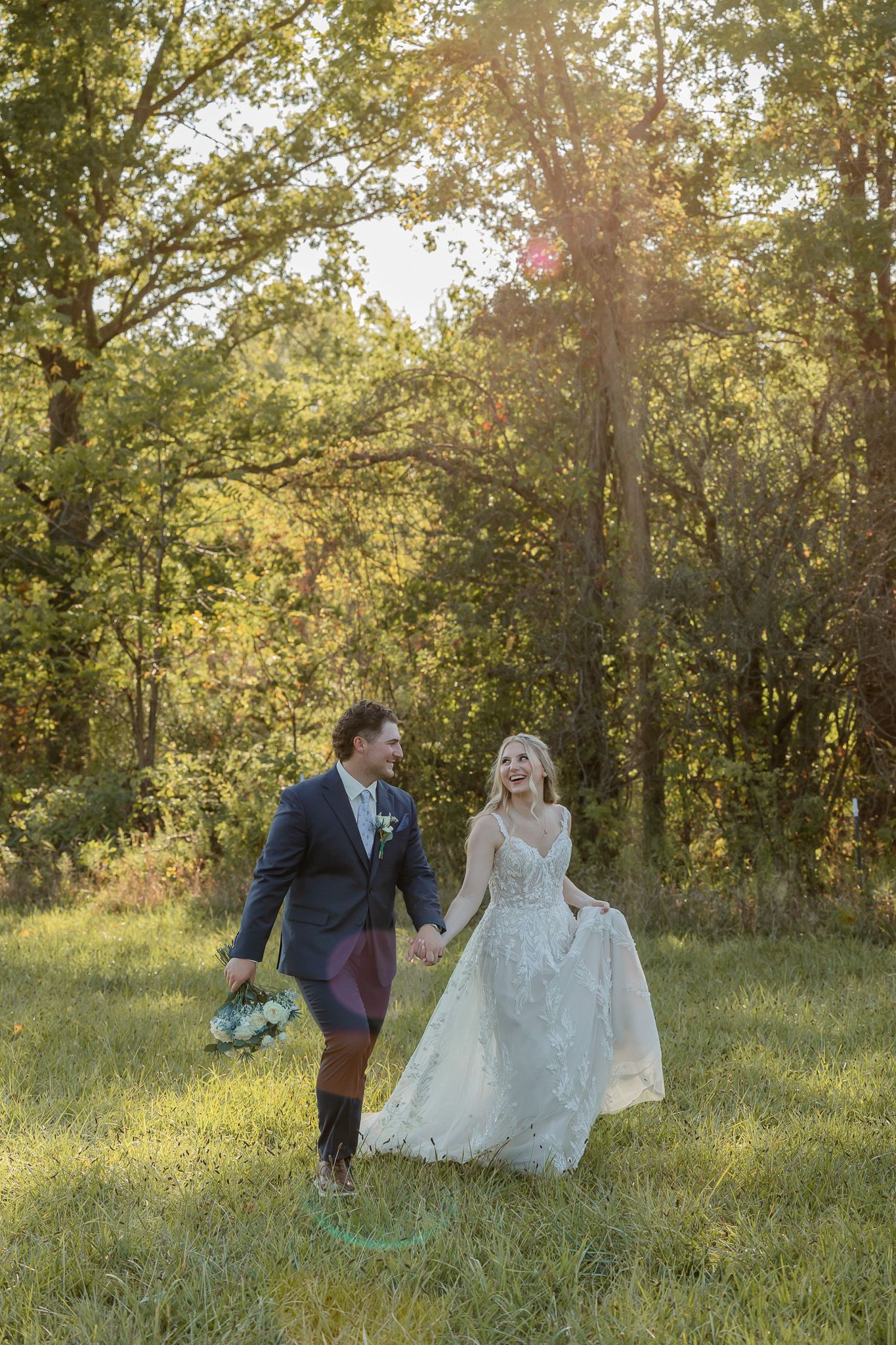 A couple walks through a sunlit meadow surrounded by trees during a wedding photoshoot at golden hour.